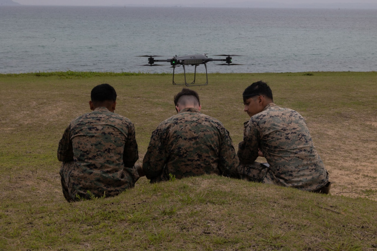 Three men in uniform sitting on grass next to a large body of water operate a remote as a drone flies above them.