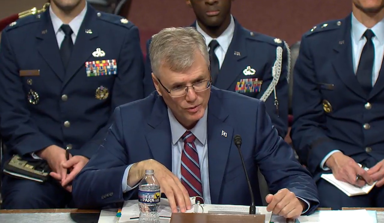 A man in a dark blue suit and glasses speaks into a microphone. Several military personnel in dress uniforms are seated in the background.