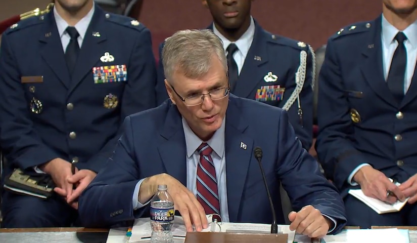 A man in a dark blue suit and glasses speaks into a microphone. Several military personnel in dress uniforms are seated in the background.