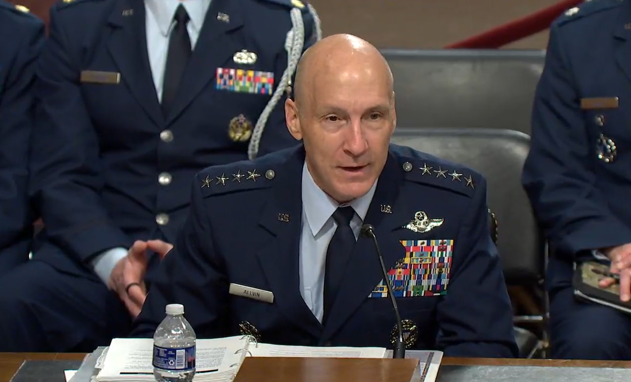 A man in a military dress uniform speaks into a microphone. Additional uniformed personnel sit in the background.