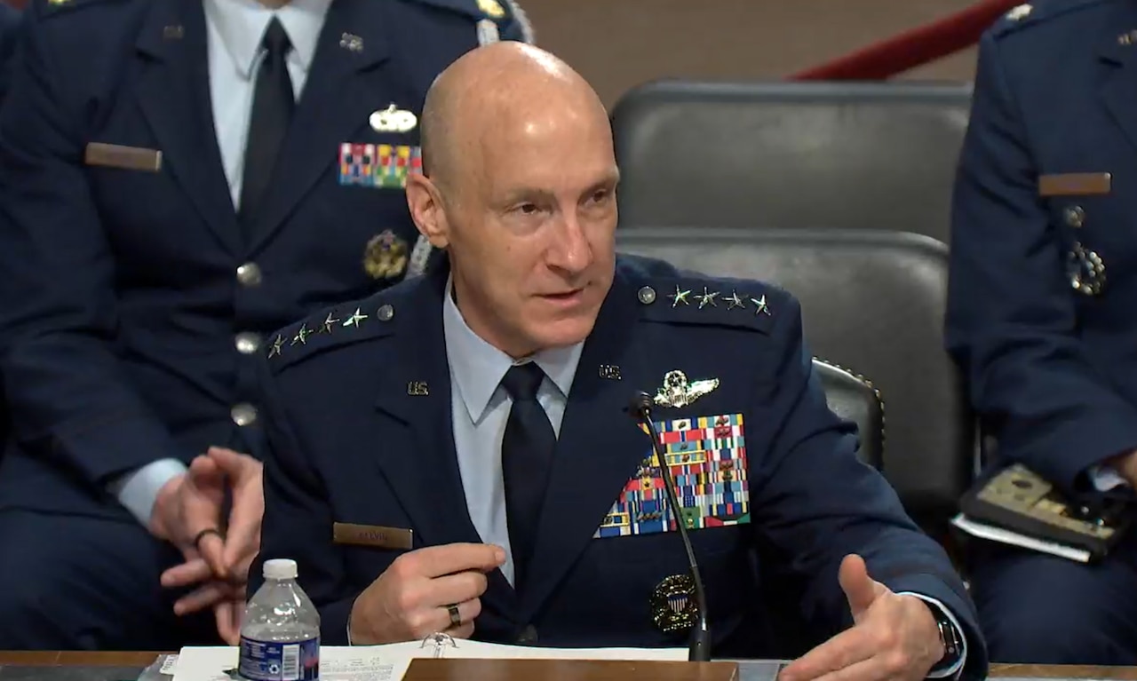 A man in a military dress uniform speaks into a microphone. Additional uniformed personnel sit in the background.