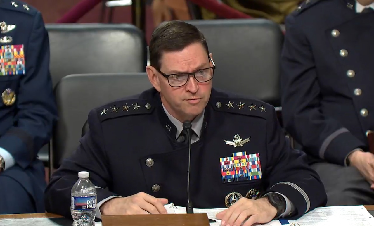A man in a military dress uniform speaks into a microphone. Additional uniformed personnel sit in the background.