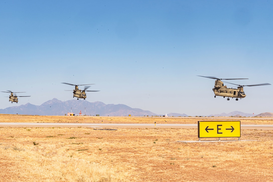 Three helicopters descend toward a rural airstrip with mountains in the distant background.