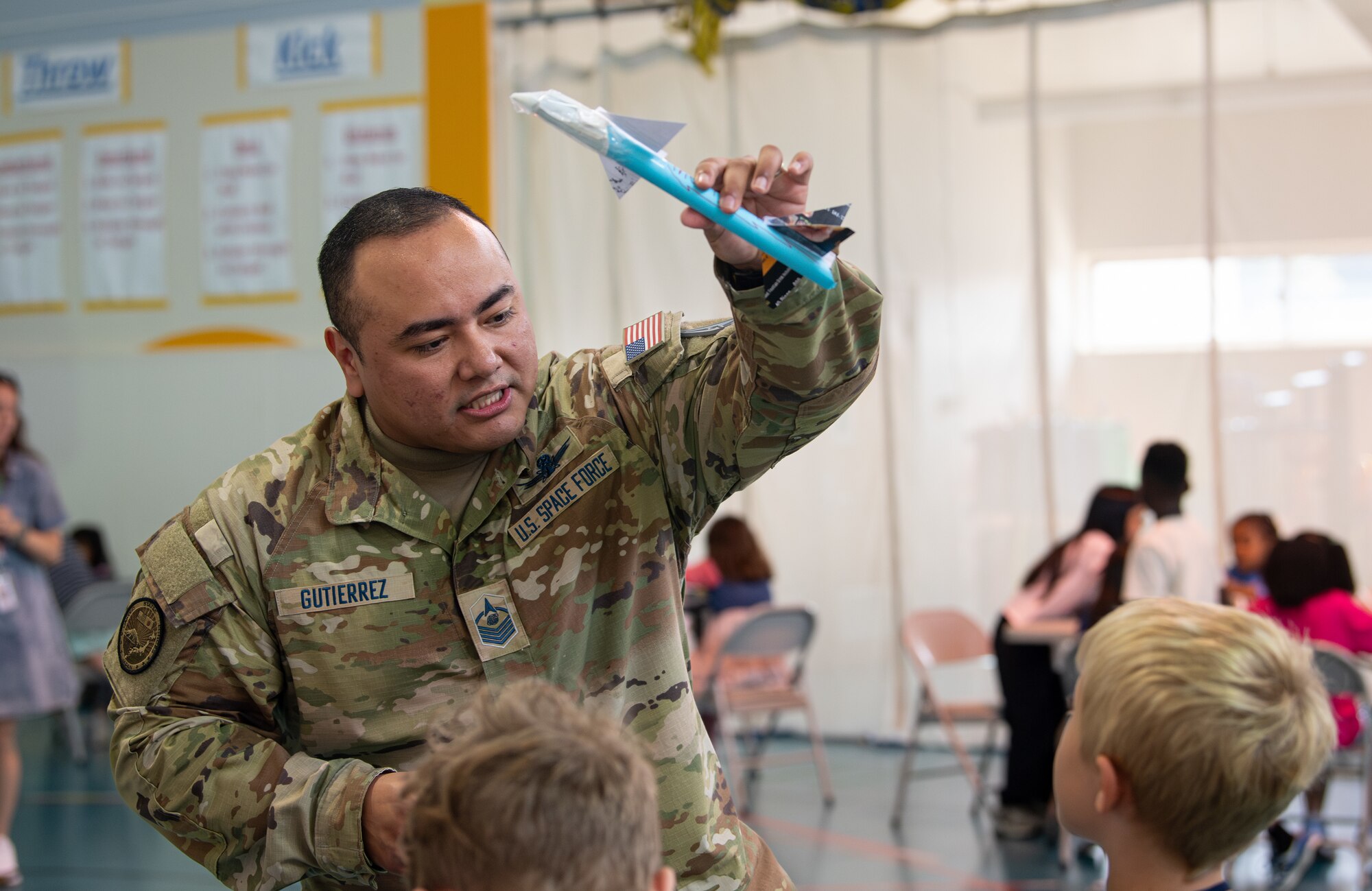 A U.S. Space Force member demonstrates a blue colored model rocket flying through the air to elementary school students.