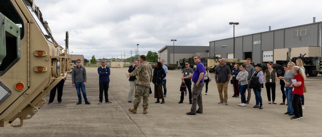 About 20 people standing outside, concrete area with warehouses behind. One man in military uniform gesturing toward a military vehicle to the left.