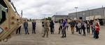 About 20 people standing outside, concrete area with warehouses behind. One man in military uniform gesturing toward a military vehicle to the left.