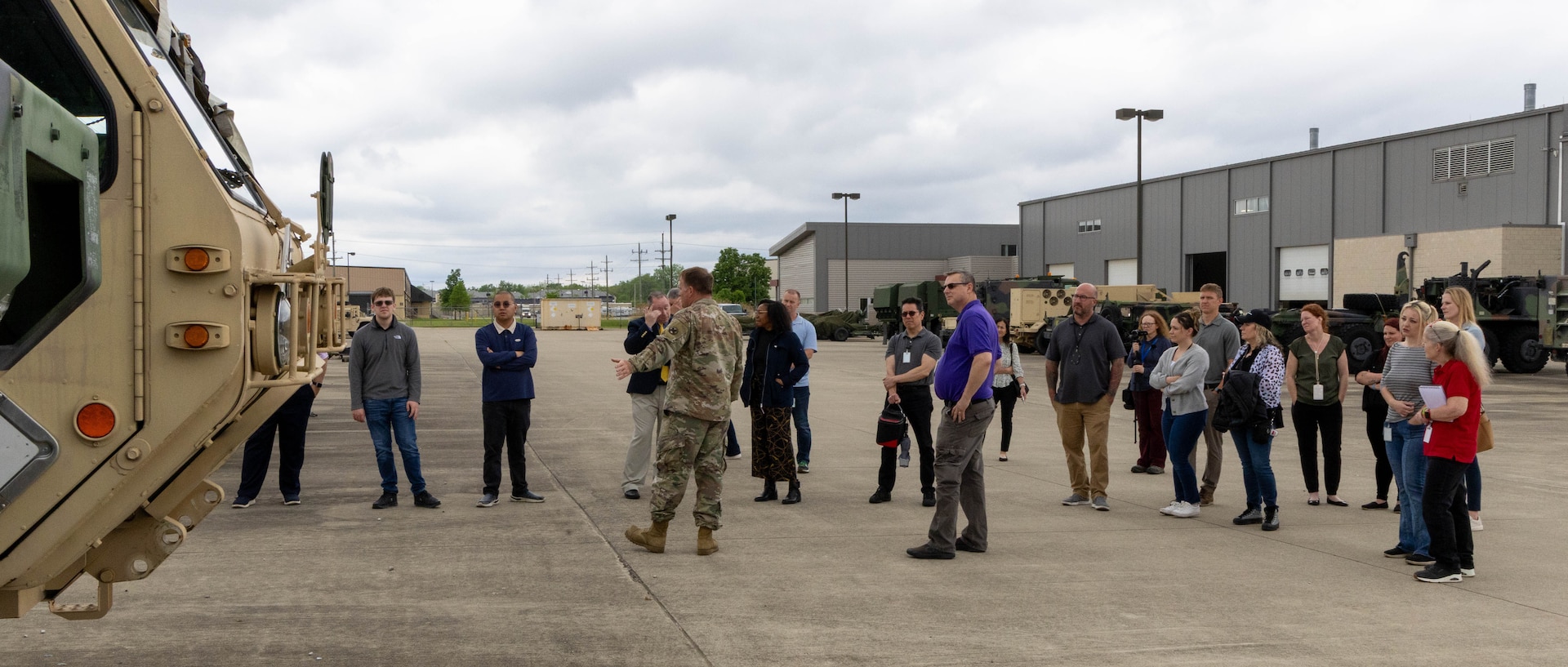 About 20 people standing outside, concrete area with warehouses behind. One man in military uniform gesturing toward a military vehicle to the left.