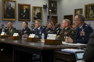Five individuals in military uniforms sit at a long table. There are painted portraits on the walls in the background.