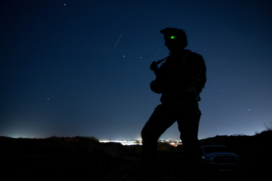 A standing soldier uses a night-vision device to look at surrounding terrain with city lights in the background.