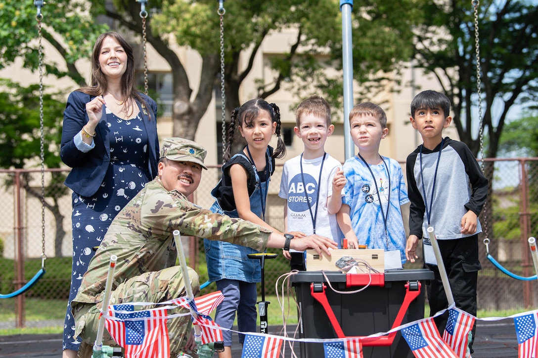 A chief engineer stands next to a guardian as they kneel next to four students in front of an American flag banner while looking up at the sky.