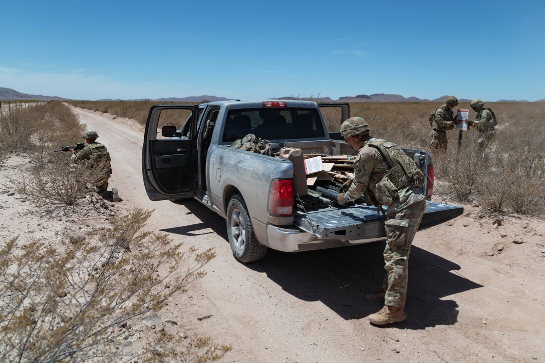 Two soldiers install a sign as another kneels and holds a rifle and a fourth pulls objects from the back of a pickup on a sandy road.