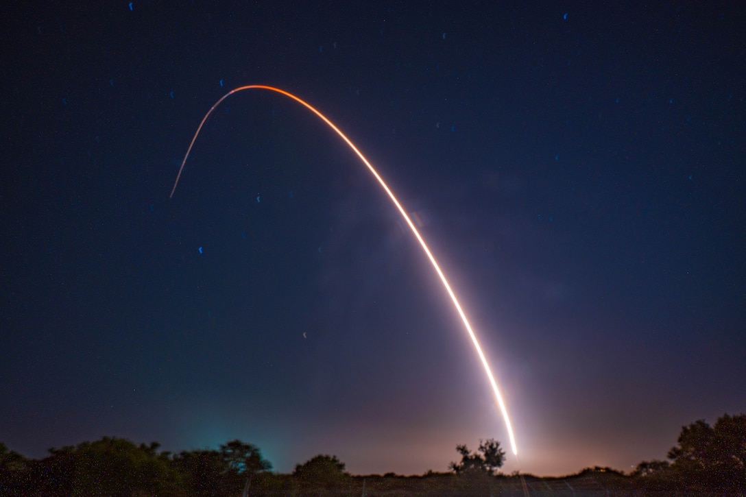 A missile leaves a light orangish streak as it launches against stars in a dark blue sky with a silhouette of a wooded area in the foreground.