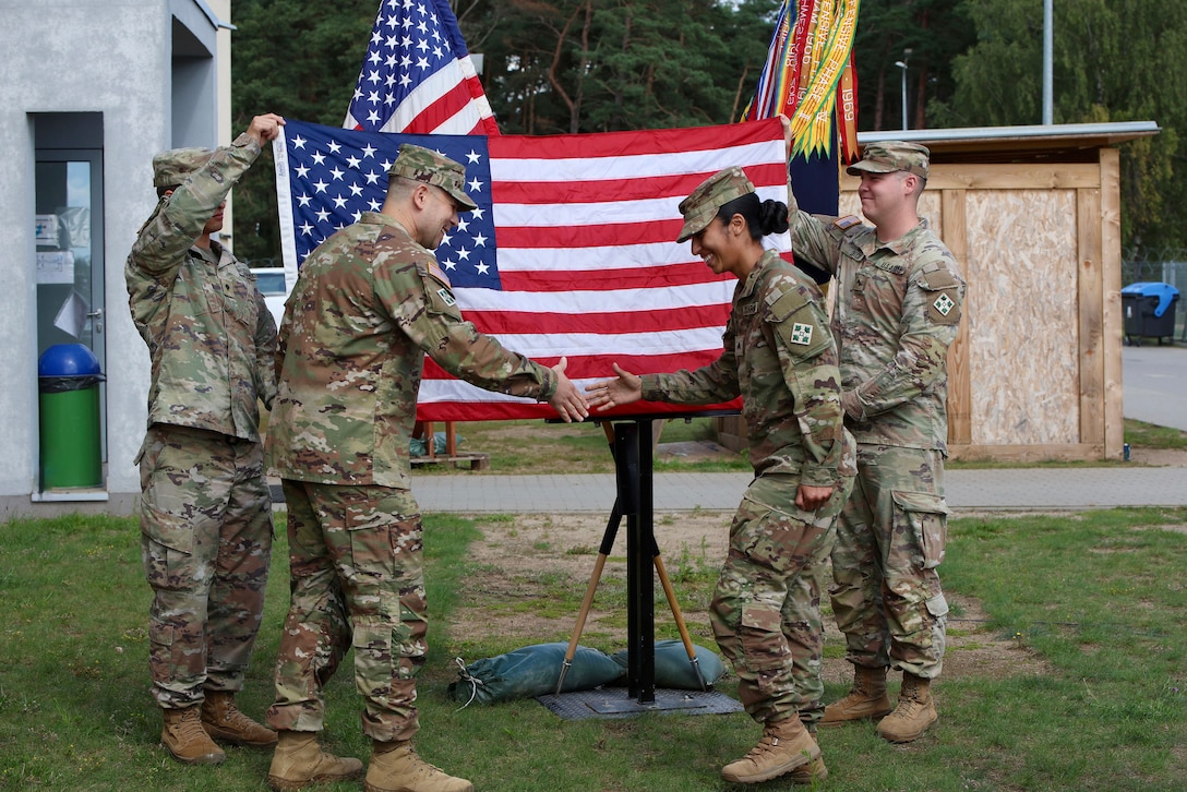 Two soldiers shake hands while standing outdoors with two other troops holding up a U.S. flag behind them.