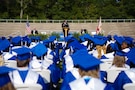 students with blue and white graduation cap and gowns sit in chairs listening to graduation speech.