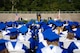 students with blue and white graduation cap and gowns sit in chairs listening to graduation speech.