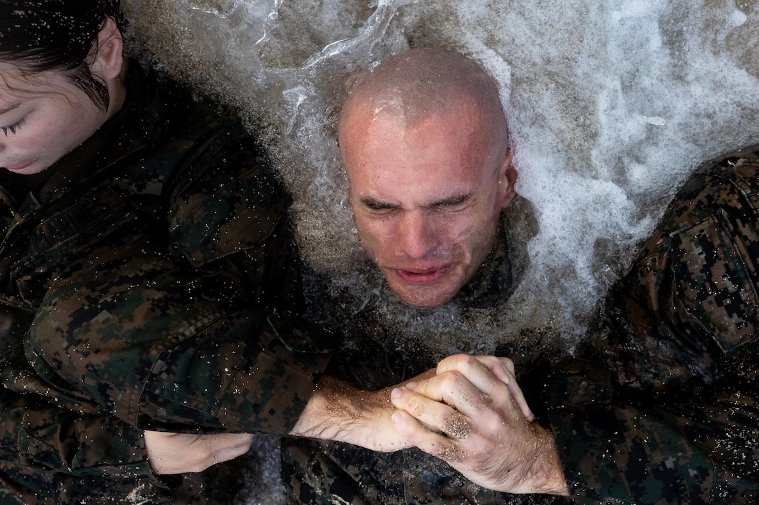 Marines in uniform and partially submerged in shallow beach water grasp arms and perform situps.