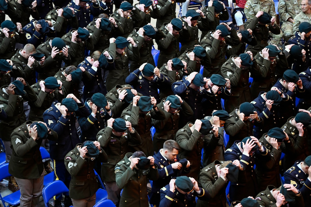 Rows of standing soldiers in dress uniforms put on green berets as others sit and watch.