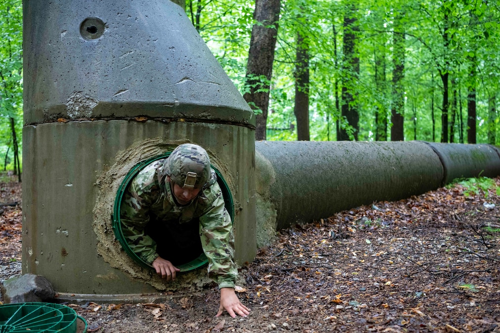 A soldier crawls out of a concrete, pipe-like tunnel in a wooded area.