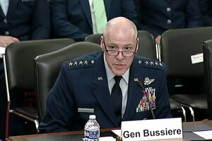 An airman in dress uniform sits at a table and speaks into a microphone.
