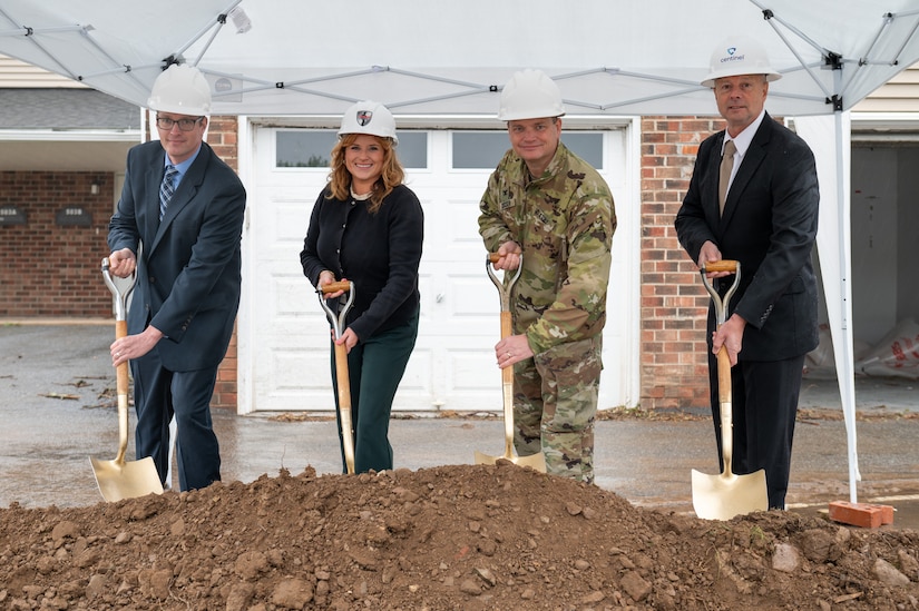 Four people all wearing white hard hats and holding gold shovels pose for a photo while digging into a large pile of dirt.