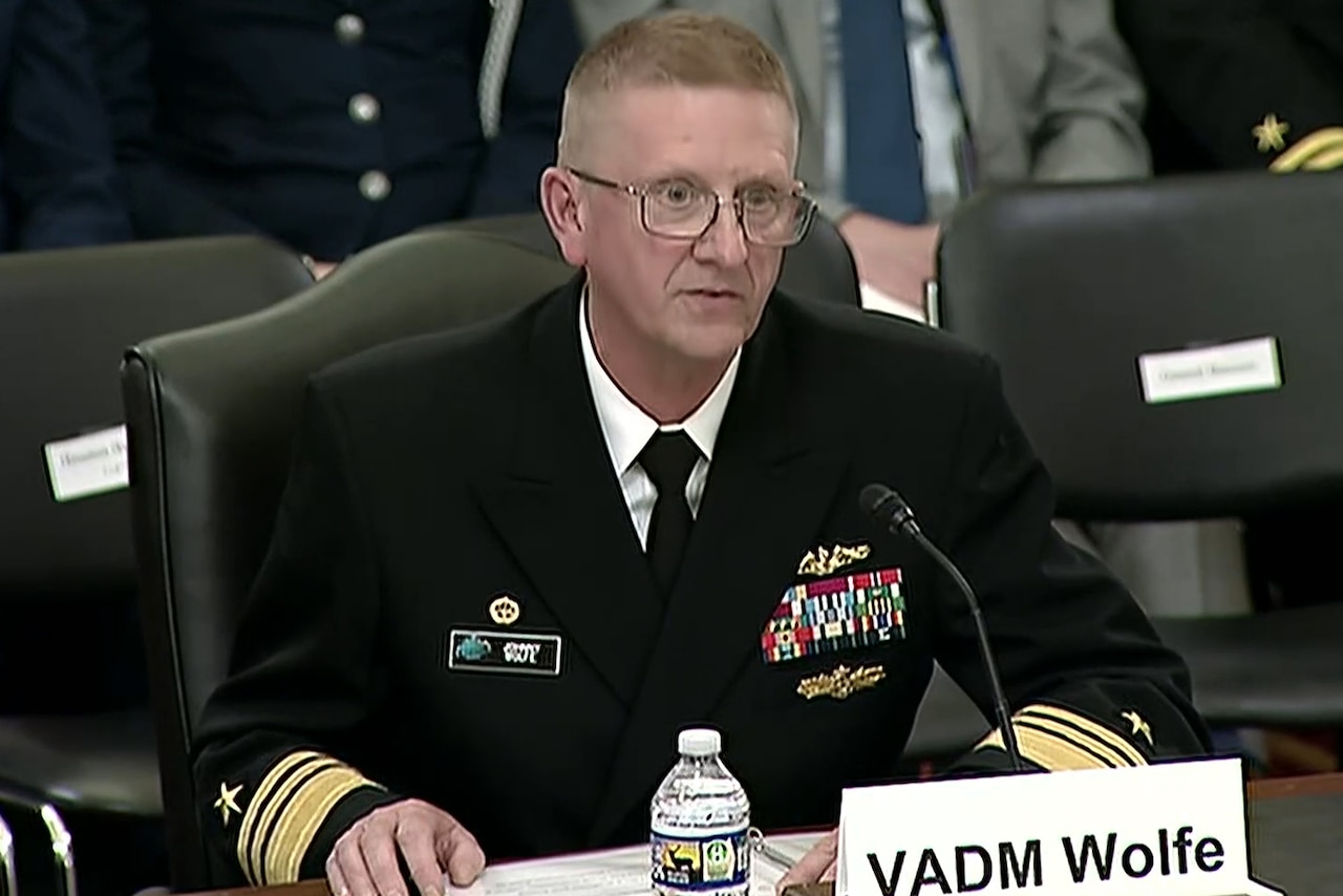 A sailor in dress uniform sits at a table and speaks into a microphone.