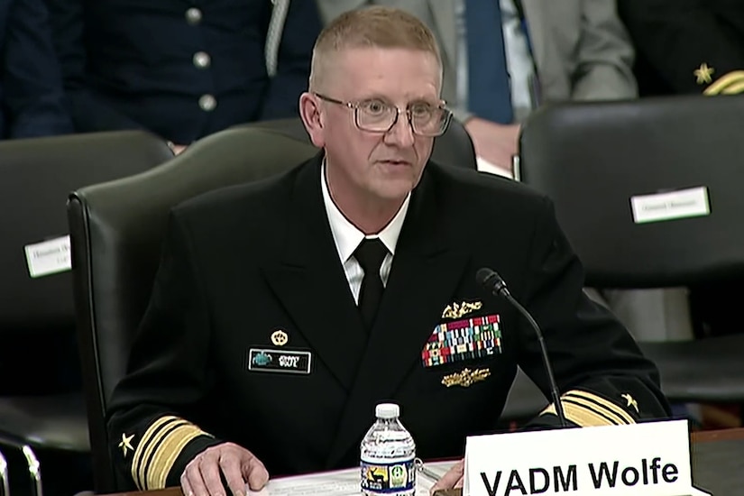A sailor in dress uniform sits at a table and speaks into a microphone.