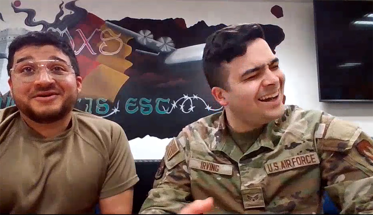 A service member wearing a camouflage uniform laughs as another man grins.