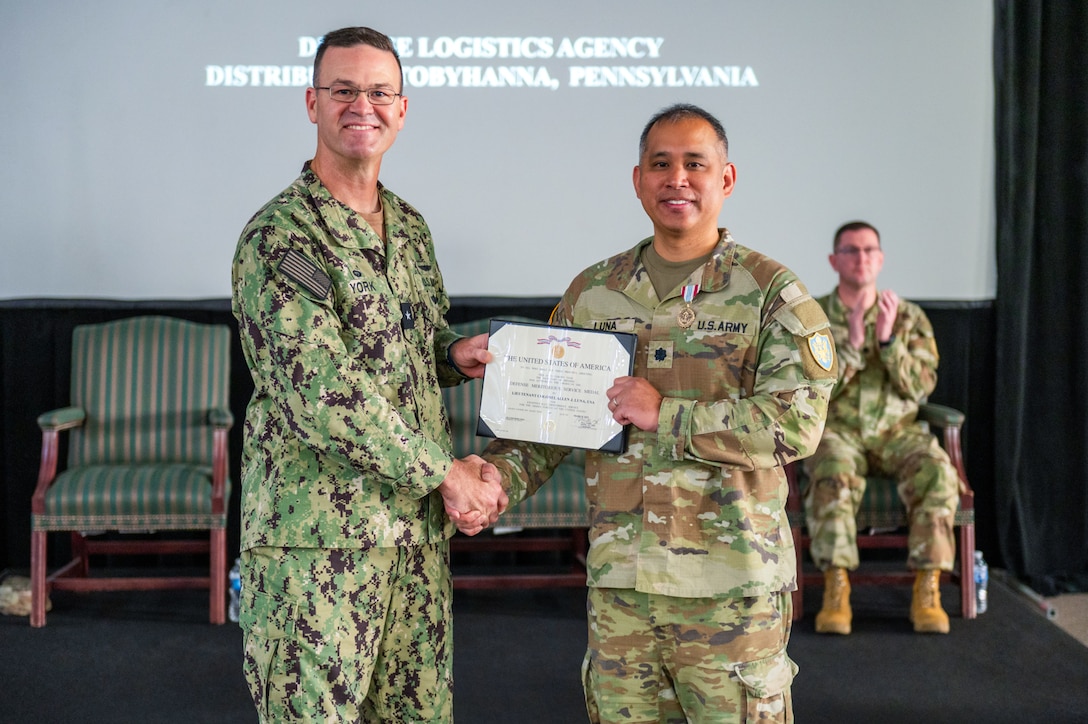 Two men wearing camouflage military uniforms stand on a stage shaking hands and holding a certificate.