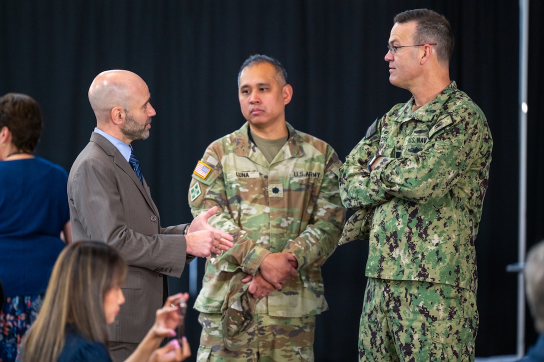 Three men stand in an auditorium speaking to one another. Two of them are wearing camouflage military uniforms.