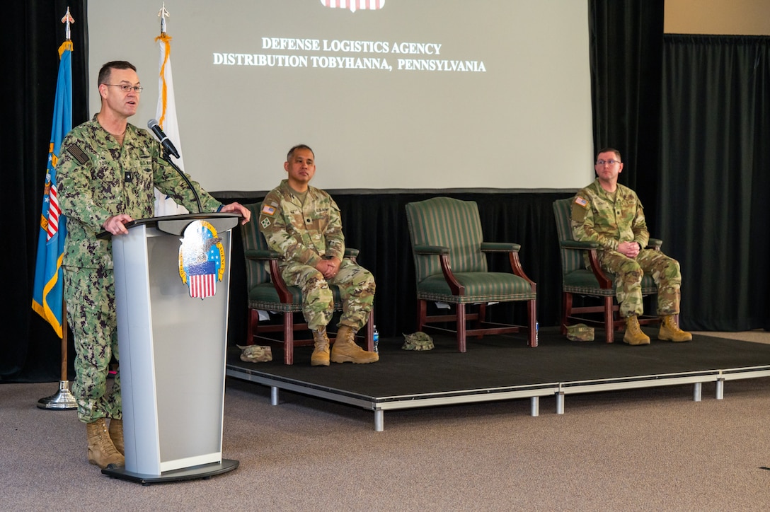 A man wearing a camouflage military uniform speaks at a lectern on a stage with flags behind him. There are two men wearing camouflage military uniforms seated on the stage to his left.