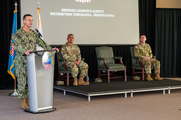 A man wearing a camouflage military uniform speaks at a lectern on a stage with flags behind him. There are two men wearing camouflage military uniforms seated on the stage to his left.