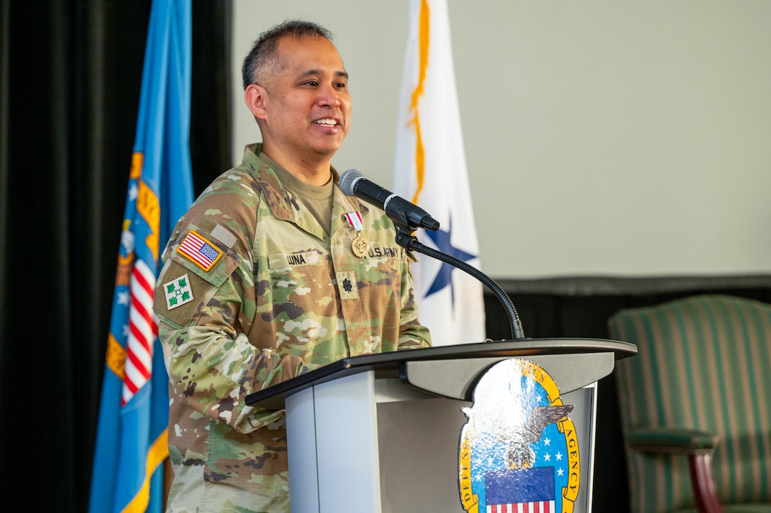 A man wearing a camouflage military uniform speaks at a lectern on a stage with flags behind him.