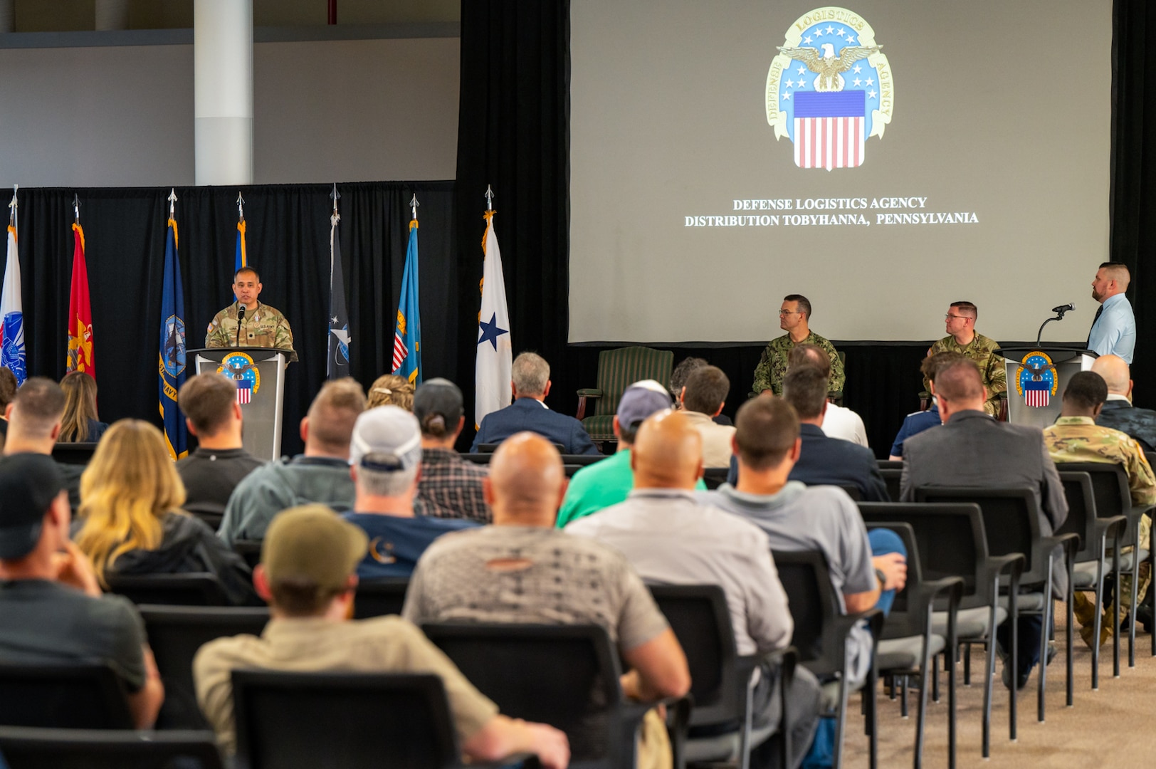 A man wearing a camouflage military uniform speaks at a lectern on a stage with flags behind him. There is a large crowd of people seated in the audience looking at him.