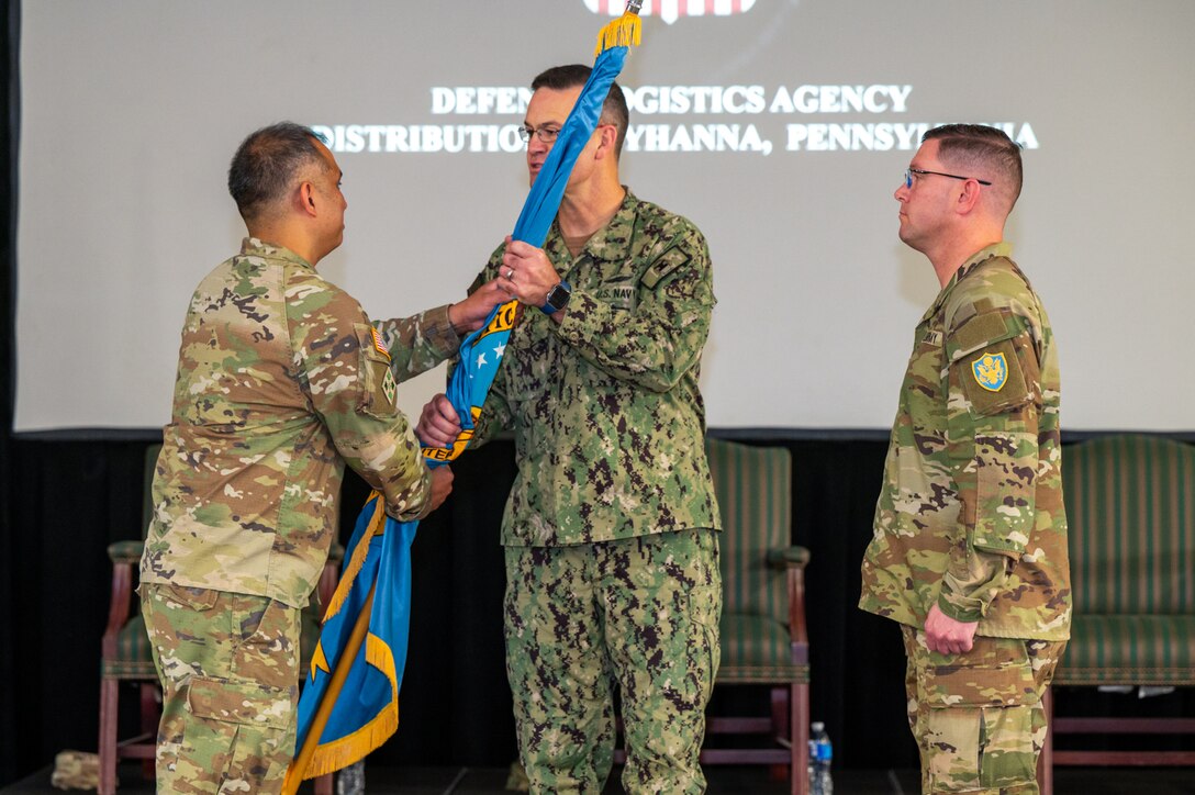 Three men wearing camouflage military uniforms stand on a stage with two passing a flag on a flagpole to one another.