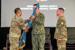 Three men wearing camouflage military uniforms stand on a stage with two passing a flag on a flagpole to one another.