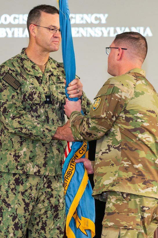 Two men wearing camouflage military uniforms pass a flag on a flagpole to one another.