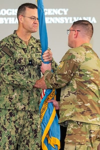 Two men wearing camouflage military uniforms pass a flag on a flagpole to one another.