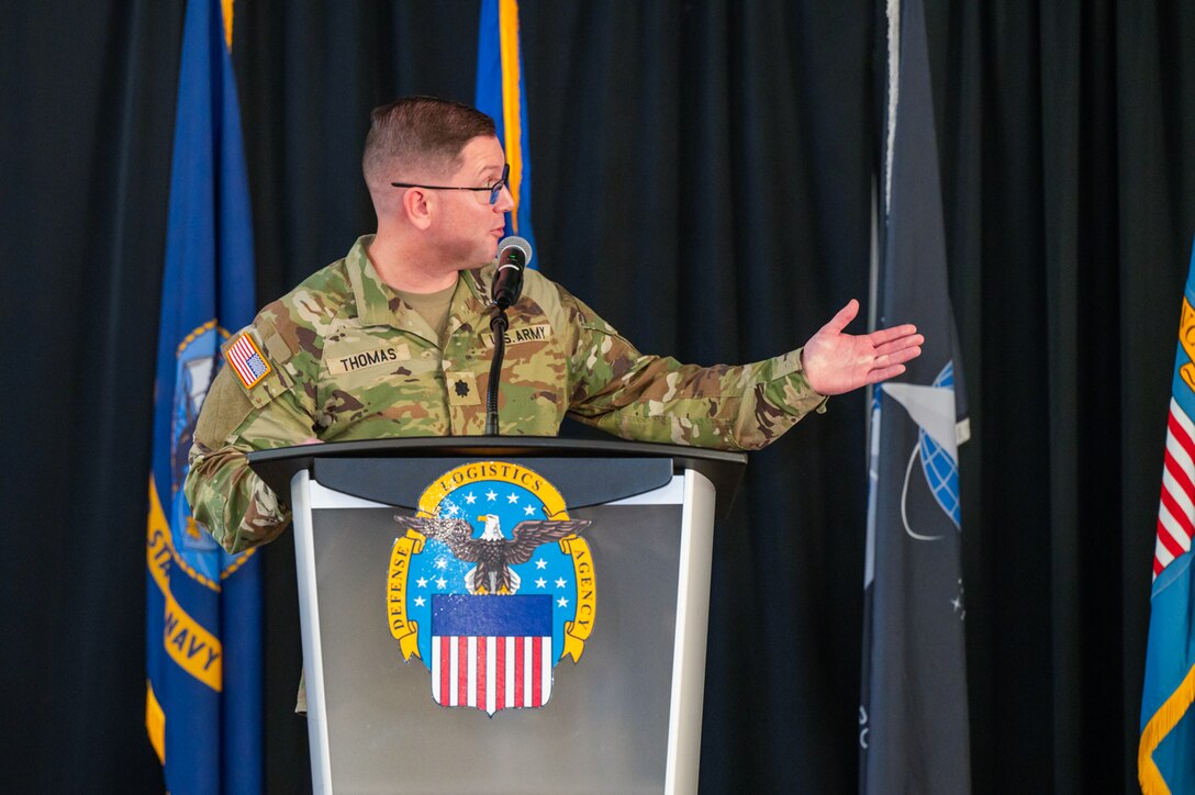 A man wearing a camouflage military uniform speaks at a lectern on a stage with flags behind him.
