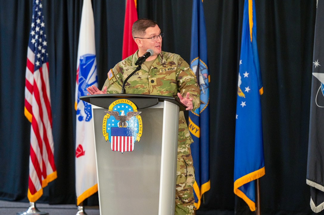 A man wearing a camouflage military uniform speaks at a lectern on a stage with flags behind him.