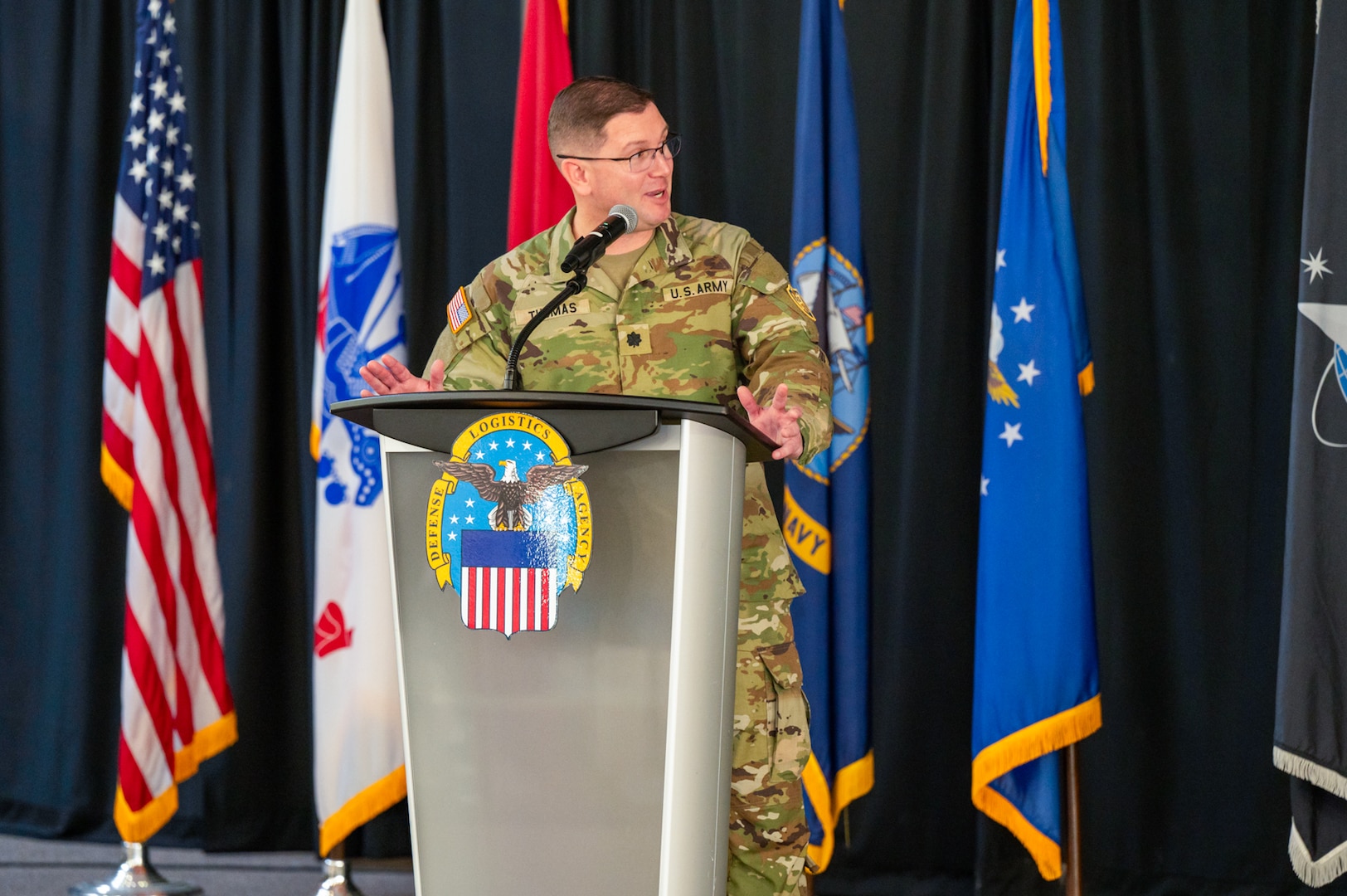 A man wearing a camouflage military uniform speaks at a lectern on a stage with flags behind him.