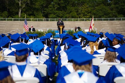 students with blue and white graduation cap and gowns sit in chairs listening to graduation speech.