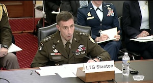A man in a military dress uniform speaks into a microphone. Two women, one in a military dress uniform and the other in business attire, sit behind him.