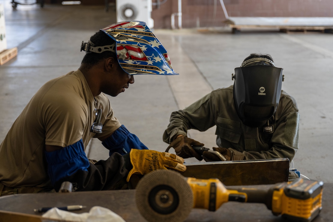 Two U.S. Air Force Airmen look over a piece of metal before performing a welding task in their shop.