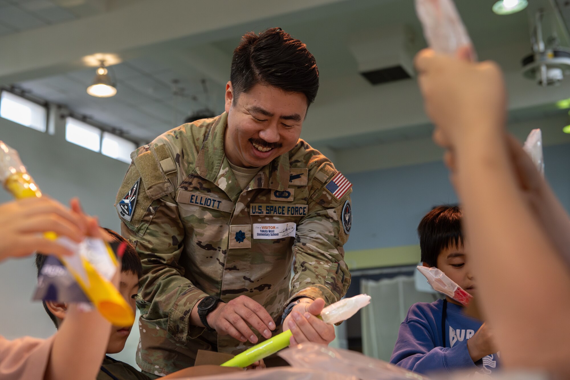 A U.S. Space Force member smiles as they hold a green model rocket while assisting students with building rockets.