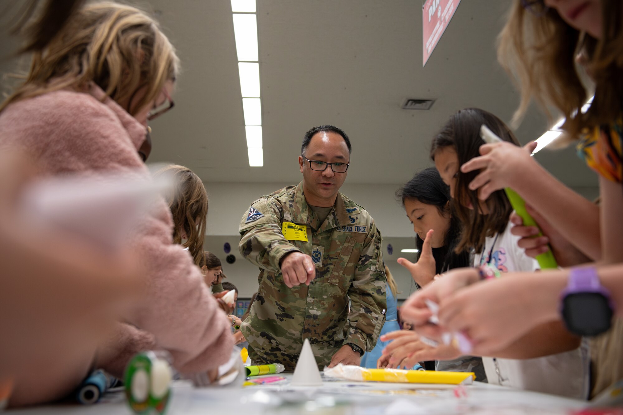 A U.S. Space Force member dressed in a military uniform is framed by the hands and arms of students as he points toward an object on a table.