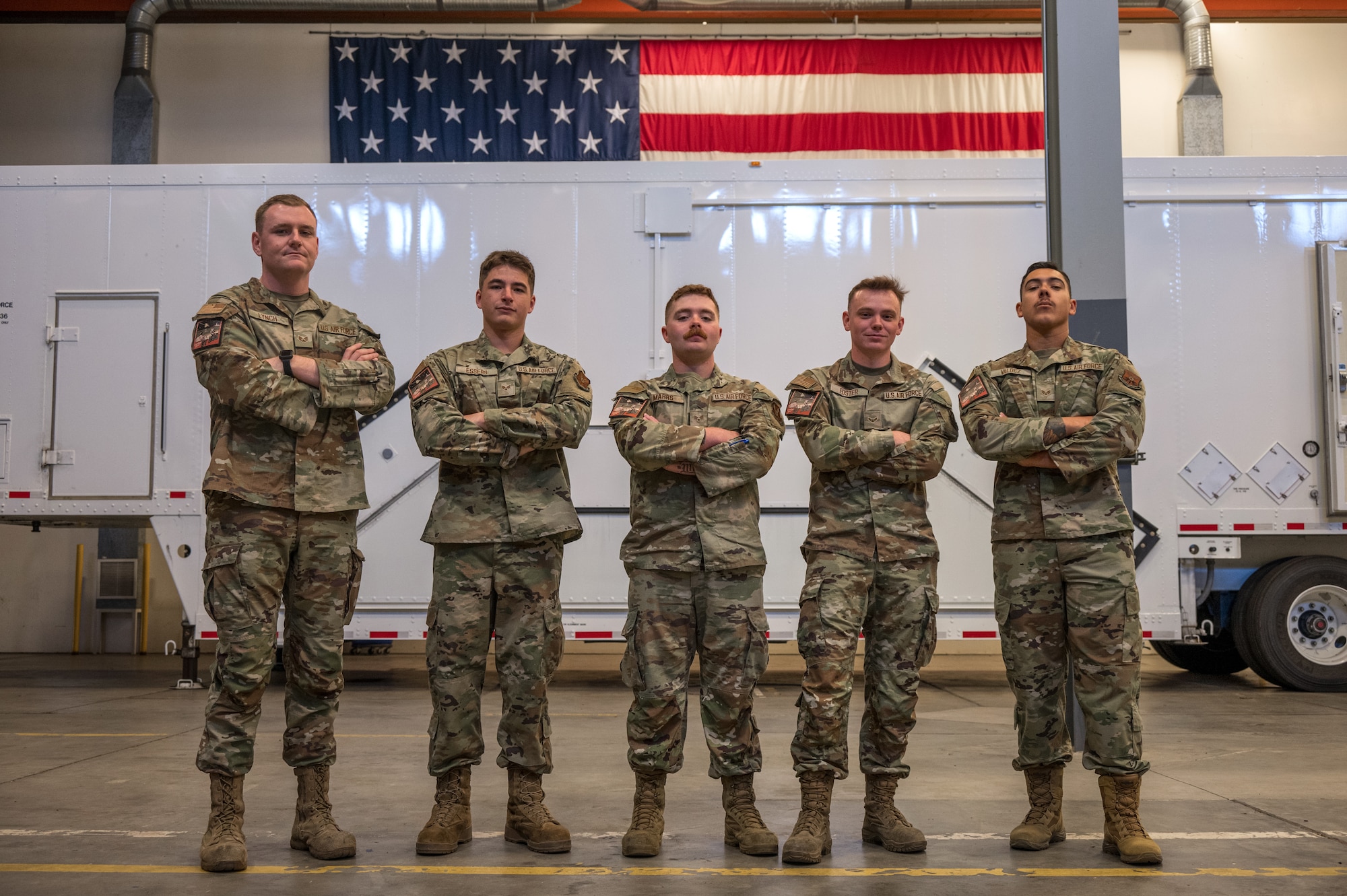 Five men in military uniform stand shoulder-to-shoulder in front of a white trailer and American flag.