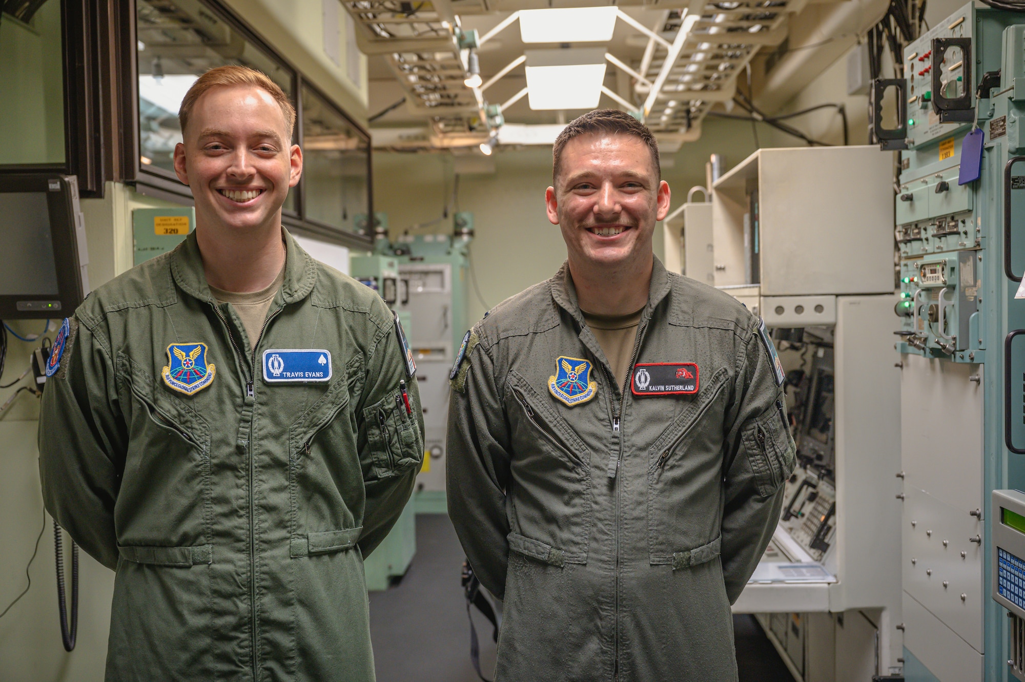 Two men in green flight suit uniforms stand shoulder to shoulder and smile at the camera.
