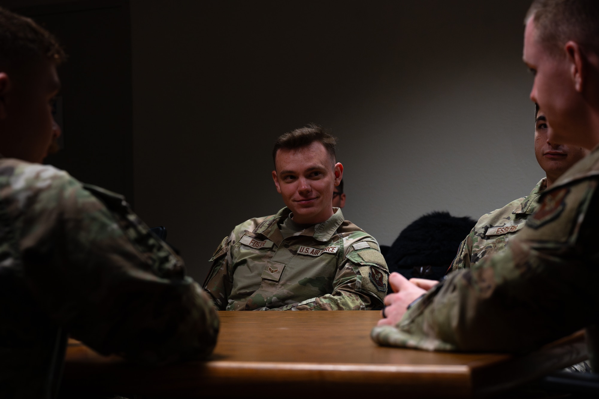 A man in military uniform sits at a conference table in a dimly lit room as he talks with other uniformed members.