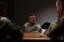 A man in military uniform sits at a conference table in a dimly lit room as he talks with other uniformed members.