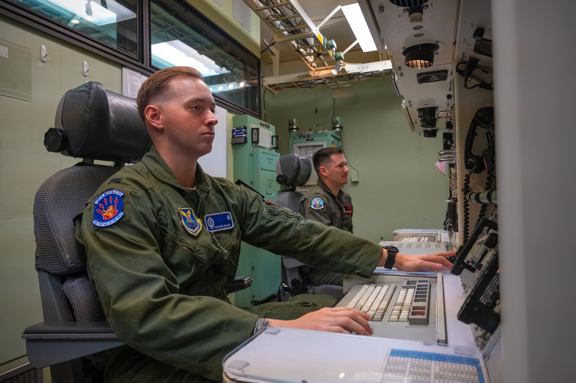 Two men in green military flight suit uniforms sit at a console table inside a launch control center capsule.